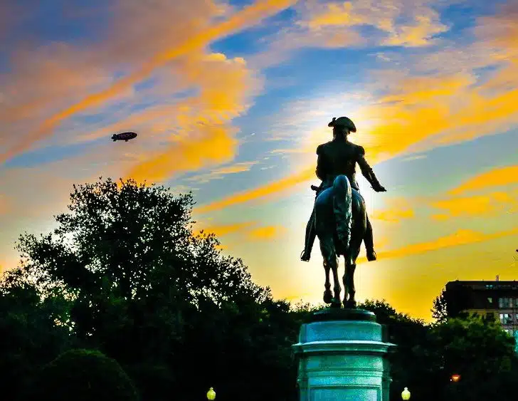 George Washington guards the Boston Public Garden entrance and watches the blimp in the pink sky.